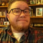 A headshot of a white middle-aged trans man with a beard and glasses